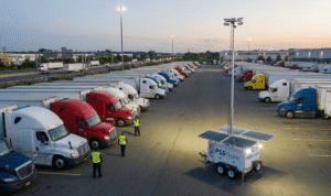 An aerial photograph taken at dusk shows a large truck stop filled with parked semi-trucks next to a multi-lane highway. In the foreground, a white PSS Onsite solar-powered mobile surveillance trailer with extended solar panels and a telescoping mast with bright lights and cameras is positioned. Three security guards wearing high-visibility vests walk among the trucks near the trailer. The lights from the surveillance unit illuminate the immediate area. In the background, traffic flows on the highway, and more trucks are parked, with some buildings and trees visible under a twilight sky.