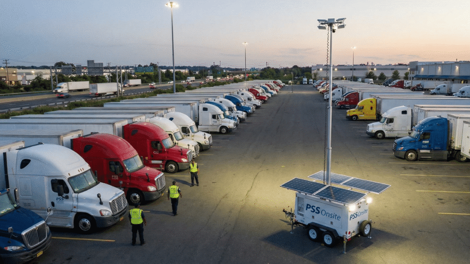 An aerial photograph taken at dusk shows a large truck stop filled with parked semi-trucks next to a multi-lane highway. In the foreground, a white PSS Onsite solar-powered mobile surveillance trailer with extended solar panels and a telescoping mast with bright lights and cameras is positioned. Three security guards wearing high-visibility vests walk among the trucks near the trailer. The lights from the surveillance unit illuminate the immediate area. In the background, traffic flows on the highway, and more trucks are parked, with some buildings and trees visible under a twilight sky.