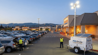 A PSS Onsite solar-powered mobile surveillance trailer with a tall mast and bright floodlights is deployed in a busy Costco parking lot at dusk. A security guard in a yellow vest stands near the trailer, while another guard speaks with a woman by a parked SUV. The trailer has solar panels on top and "PSS Onsite" and "Solar Powered Mobile Surveillance" logos. The parking lot is filled with cars, and other stores, including a Verizon, are visible in the background under a darkening sky.