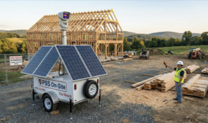 A Solar portable construction security unit with 'Fire Watch' branding stands on a rural construction site next to a timber-framed building, with a worker holding a device and lumber in the foreground